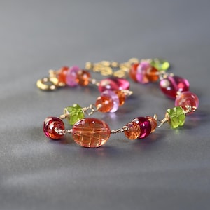 May include: A bracelet featuring a series of faceted, multi-colored beads in shades of pink, orange, red, and lime green, connected by thin gold-toned wire links. The beads vary in shape, including round, oval, and square, with a gold-toned clasp.