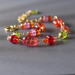 May include: A bracelet featuring a series of multi-colored translucent beads in shades of pink, orange, red, and lime green, connected by thin gold-toned wire links. The central bead is a large, oval-shaped pink piece with a carved grid pattern.