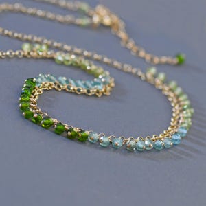 May include: A delicate gold necklace featuring a gradient of green and blue gemstones. The necklace has a fine chain and is adorned with small, round beads that transition from deep green to light blue. The design is elegant and suitable for various occasions.