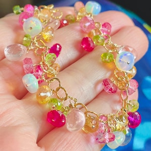 Può includere: Un delicato braccialetto d'oro ornato da una serie di pietre preziose colorate. Il braccialetto presenta una varietà di forme e dimensioni, tra cui pietre rotonde, ovali e sfaccettate in tonalità rosa, verde, giallo e trasparente. Le pietre preziose sono collegate con maglie di catena d'oro.