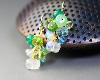 Puede incluir: Un par de pendientes colgantes con un racimo de piedras preciosas verdes, azules y blancas. Los pendientes están hechos con alambre de oro y tienen un estilo delicado y bohemio.