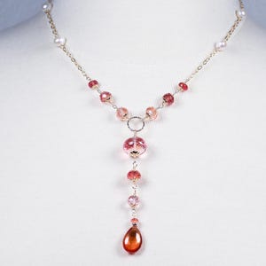 Op de afbeelding: Een delicate ketting met een gouden ketting, roze en heldere kralen en een Y-vormig ontwerp. Het heeft een grote roze kristallen hanger en een druppelvormige amberkleurige hanger. Kleine parels bevinden zich aan de bovenkant van de ketting.