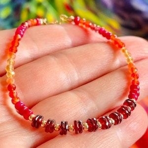 May include: A beaded bracelet featuring a gradient of small, faceted gemstones in shades of red, orange, and yellow. The design incorporates round, polished gold-tone metal beads interspersed between the faceted stones, creating a vibrant, multi-colored accessory with a delicate, circular structure.