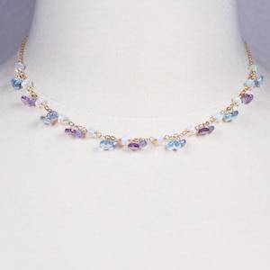 May include: Delicate gold-tone necklace featuring a series of small, colorful beads. The necklace has a delicate chain with clusters of blue, purple, and white beads. The beads are arranged in a cascading pattern, creating a subtle, elegant design.