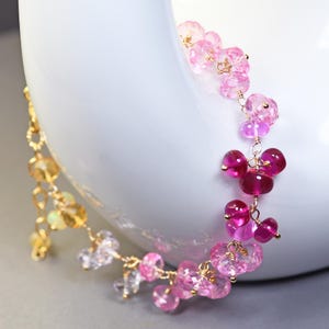 May include: A delicate gold chain bracelet adorned with clusters of pink and clear gemstones. The bracelet features varying shades of pink, from light to deep magenta, and is displayed against a white background.