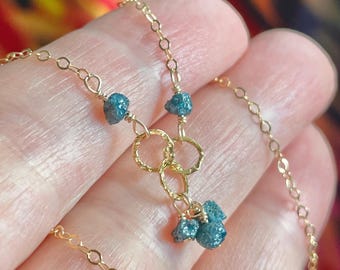 May include: A delicate gold necklace featuring three clusters of rough, blue-green gemstones. The necklace has a chain with small, linked circles and three textured gold rings. The gemstones are wire-wrapped to the chain.