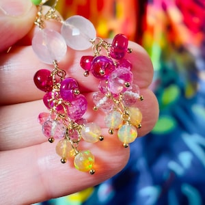 May include: Close-up of gold-toned earrings with clusters of colorful gemstones. The earrings feature pink, red, and yellow-orange faceted and round stones. The gemstones are suspended from delicate gold chains.