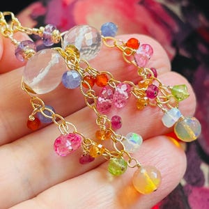 May include: A gold chain earring featuring a cluster of multi-colored faceted gemstones including clear quartz, pink tourmaline, orange carnelian, blue tanzanite, green peridot, and iridescent opals, all suspended from delicate gold wire links.