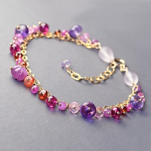 May include: A gold chain bracelet with a gradient of pink, red, and purple gemstones. The bracelet features a variety of shapes and sizes of gemstones, including round, oval, and faceted.