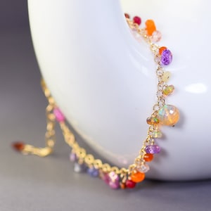May include: A gold chain bracelet with a variety of colorful gemstones, including orange, red, pink, purple, and white. The bracelet is on a white surface.