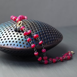 May include: A double strand bracelet with pink gemstone beads and a black chain. The bracelet has a small gold charm with black gemstones.