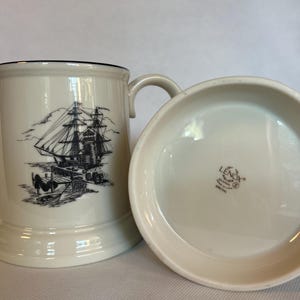 Può includere: Set tazza e piattino Lenox color crema. La tazza presenta un'illustrazione nera di una nave alta, mentre il piattino è semplice. La tazza ha un bordo nero e un manico curvo. Il logo Lenox è visibile sul piattino.