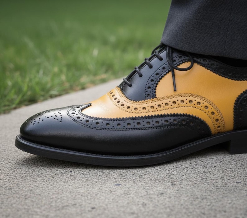 K&ouml;nnte beinhalten: Nahaufnahme eines zweifarbigen Lederschuhs. Der Schuh hat eine schwarze Basis mit einem senfgelben Oberteil. Der Schuh hat dekorative Perforationen und schwarze Schn&uuml;rsenkel. Der Schuh steht auf einer Betonoberfl&auml;che.