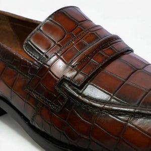 Op de afbeelding: Close-up van een bruine leren loafer met een krokodillenleertextuur. De schoen heeft een band over de bovenkant en een zwarte zool. Het leer heeft een glanzende afwerking.