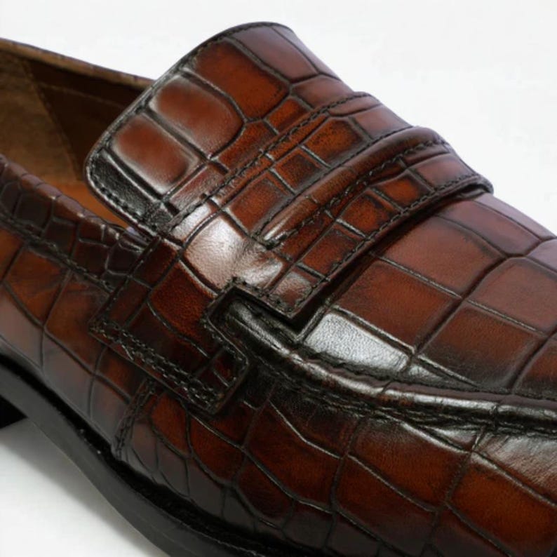 Op de afbeelding: Close-up van een bruine leren loafer met een krokodillenleertextuur. De schoen heeft een band over de bovenkant en een zwarte zool. Het leer heeft een glanzende afwerking.