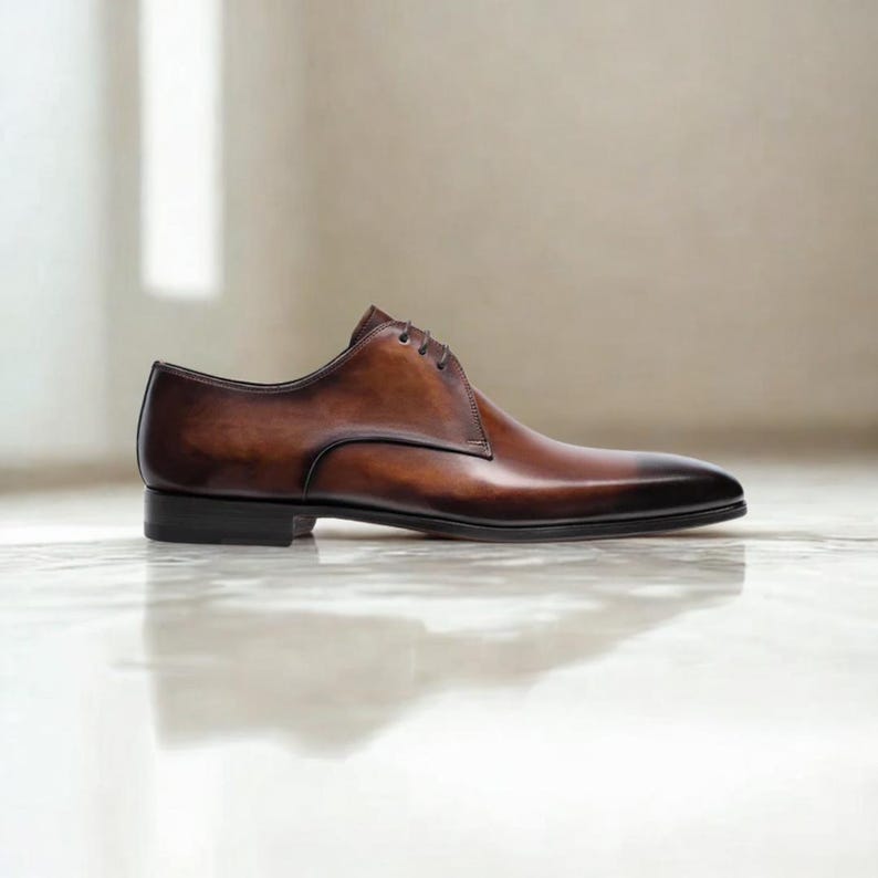 May include: A single brown leather dress shoe with a pointed toe and a low heel. The shoe has a polished finish and is laced up. The background is a neutral color, and the shoe is sitting on a reflective surface.