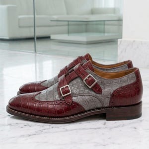 K&ouml;nnte beinhalten: Ein Paar Doppel-Monk-Schuhe in zweifarbigem Design aus dunkelburgunderfarbenem und grauem Leder mit Krokodilpr&auml;gung. Die Schuhe verf&uuml;gen &uuml;ber silberfarbene Metallschnallen, eine Fl&uuml;gelkappe mit Brogue-Verzierungen und einen flachen Absatz.