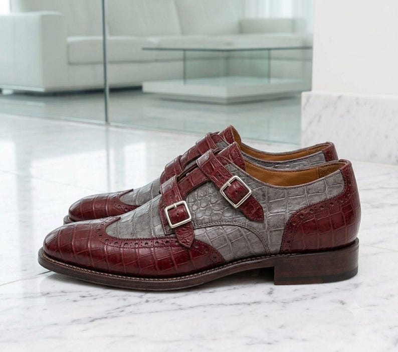 K&ouml;nnte beinhalten: Ein Paar Doppel-Monk-Schuhe in zweifarbigem Design aus dunkelburgunderfarbenem und grauem Leder mit Krokodilpr&auml;gung. Die Schuhe verf&uuml;gen &uuml;ber silberfarbene Metallschnallen, eine Fl&uuml;gelkappe mit Brogue-Verzierungen und einen flachen Absatz.