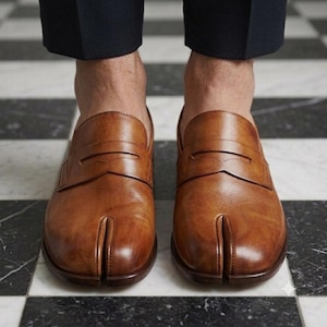 Op de afbeelding: Een paar lichtbruine leren loafers met een opvallend ontwerp met gespleten neus aan de voorzijde. De schoenen hebben een klassieke penny loafer-band over de wreef en een gladde, gepolijste afwerking met een lage hak en een ronde vorm.