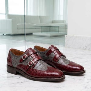K&ouml;nnte beinhalten: Ein Paar Doppel-Monk-Schuhe mit zweifarbigem Design aus dunkelburgunderfarbenem und grauem Leder in Krokodilpr&auml;gung. Die Schuhe haben silberfarbene Schnallen, eine Fl&uuml;gelkappe mit Brogue-Verzierungen und einen Absatz aus braunem Leder.