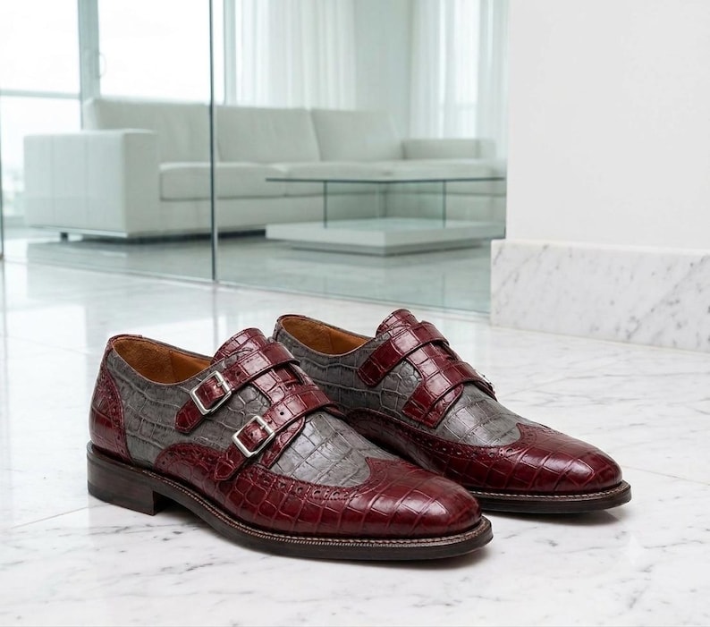 K&ouml;nnte beinhalten: Ein Paar Doppel-Monk-Schuhe mit zweifarbigem Design aus dunkelburgunderfarbenem und grauem Leder in Krokodilpr&auml;gung. Die Schuhe haben silberfarbene Schnallen, eine Fl&uuml;gelkappe mit Brogue-Verzierungen und einen Absatz aus braunem Leder.