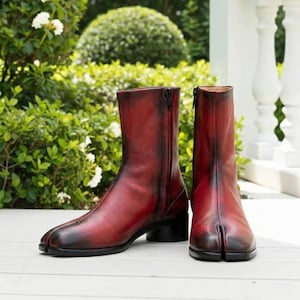 May include: A pair of red leather ankle boots with a split-toe design. The boots have a side zipper and a small black heel. The leather has a distressed appearance, with darker shading near the toe and heel.
