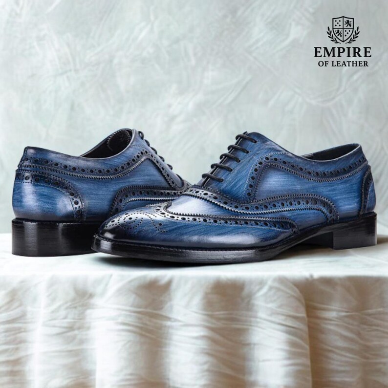 May include: A pair of blue leather brogue shoes with black soles and laces. The shoes have decorative perforations and are displayed on a white surface. The brand logo "EMPIRE OF LEATHER" is visible.