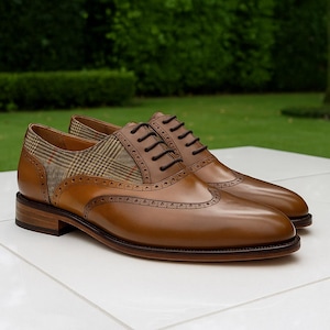 Op de afbeelding: Een paar bruine leren schoenen met wingtip-design en een geruit stoffen accent. De schoenen hebben donkerbruine veters en een gepolijste afwerking. De achtergrond is een vage groene buitenomgeving.