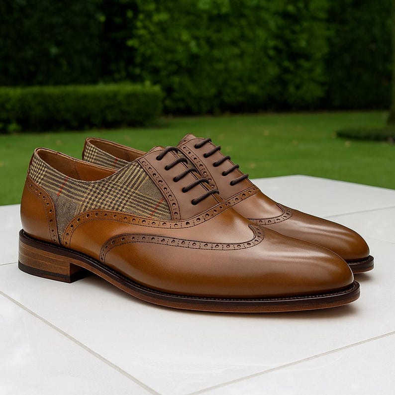 Op de afbeelding: Een paar bruine leren schoenen met wingtip-design en een geruit stoffen accent. De schoenen hebben donkerbruine veters en een gepolijste afwerking. De achtergrond is een vage groene buitenomgeving.