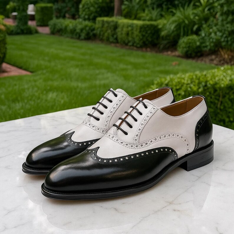 Puede incluir: Un par de zapatos de vestir de cuero de dos tonos. Los zapatos son negros y blancos con cordones negros y un dise&ntilde;o cl&aacute;sico de ala. Los zapatos se exhiben sobre una superficie de m&aacute;rmol con un fondo verde.