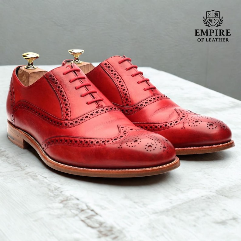 May include: A pair of red leather brogue shoes with intricate perforations and red laces. The shoes have a classic wingtip design and are displayed with wooden shoe trees. The logo "EMPIRE OF LEATHER" is visible in the background.