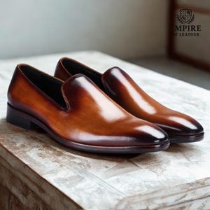 Op de afbeelding: Een paar bruin lederen instappers met een glanzende afwerking. De schoenen hebben een klassiek ontwerp met een lage hak en een ronde neus. Het Empire of Leather-logo is zichtbaar op de achtergrond.