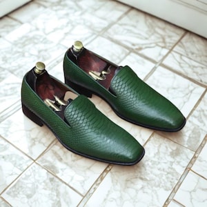 Peut inclure: Paire de mocassins en cuir vert avec un motif texturé en forme de peau de serpent. Les chaussures ont un bout carré et un petit talon. Des embauchoirs sont à l'intérieur. Les mocassins sont présentés sur une surface carrelée.