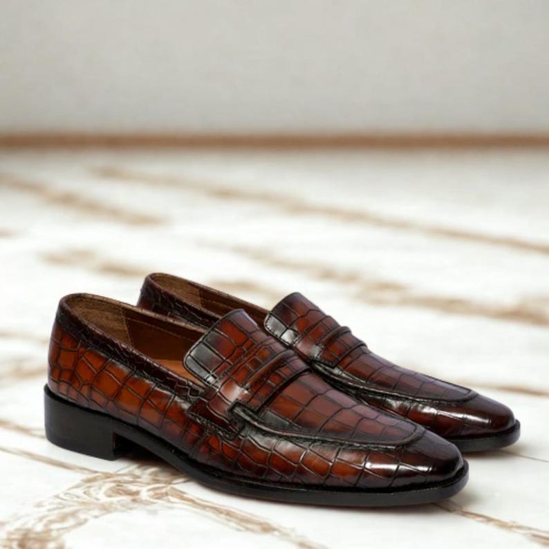 Op de afbeelding: Een paar bruine leren loafers met een krokodillenleertextuur. De schoenen hebben een klassiek ontwerp met een lage hak en een instapmodel. Het leer heeft een glanzende afwerking en een donkerbruine kleur.