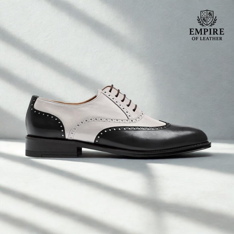 Puede incluir: Un zapato Oxford de cuero de dos tonos. El zapato presenta una puntera y un tal&oacute;n negros, con una parte superior blanca. El zapato tiene un dise&ntilde;o cl&aacute;sico de punta de ala con perforaciones decorativas y cordones marrones. El logotipo de Empire of Leather est&aacute; en la esquina superior derecha.
