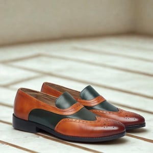 Op de afbeelding: Tweekleurige leren loafers in bruin en donkergroen. De schoenen hebben decoratieve perforaties op de neus en zwarte zolen. Ze staan op een lichtgekleurd houten oppervlak. Klassiek en stijlvol ontwerp.