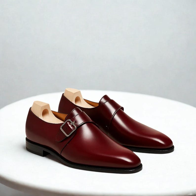 May include: A pair of burgundy leather monk strap shoes with silver buckles. The shoes have a sleek, polished appearance and are displayed on a white surface. Wooden shoe trees are inserted inside the shoes.