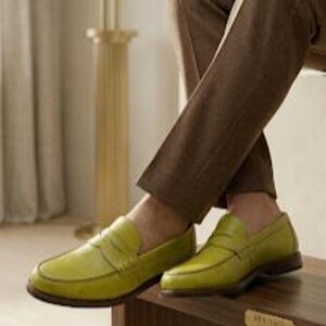 Puede incluir: Un par de mocasines de cuero verde-amarillo brillante con detalle de correa de centavo. Los zapatos se exhiben sobre una superficie de madera, combinados con pantalones marrones. Los mocasines tienen un aspecto cl&aacute;sico y pulido.