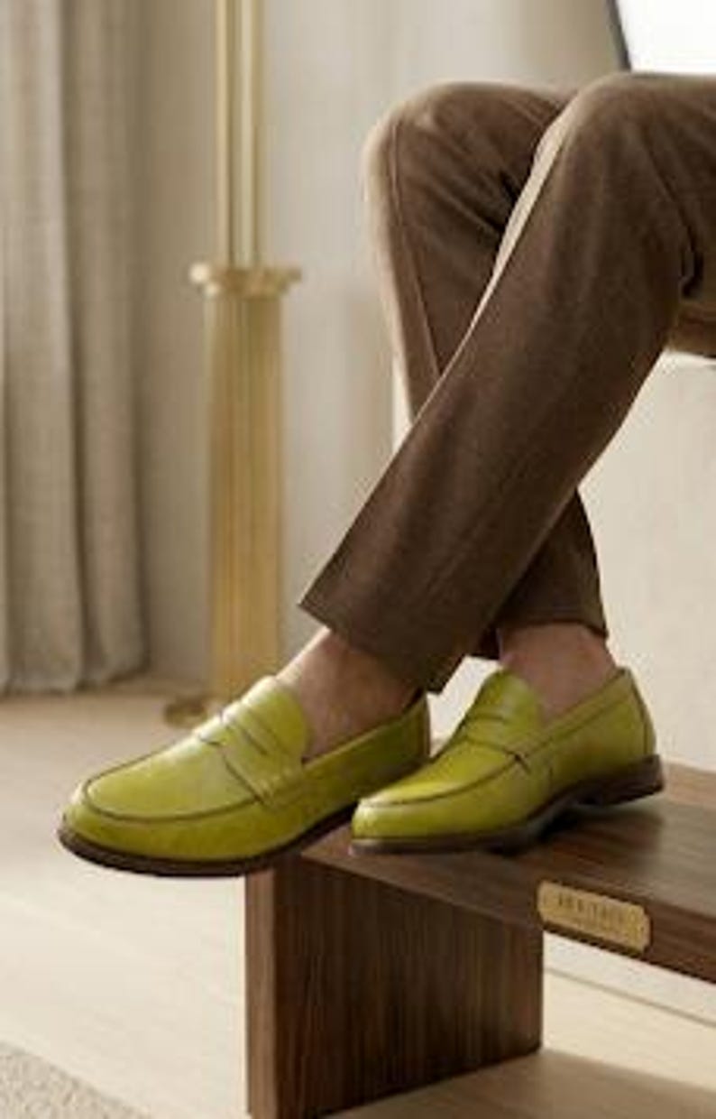 Puede incluir: Un par de mocasines de cuero verde-amarillo brillante con detalle de correa de centavo. Los zapatos se exhiben sobre una superficie de madera, combinados con pantalones marrones. Los mocasines tienen un aspecto cl&aacute;sico y pulido.