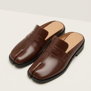 May include: A pair of dark brown leather backless loafers featuring a distinctive split-toe design at the front. These slip-on shoes have a smooth, polished finish, a traditional penny loafer strap across the vamp, and a black rubber sole.