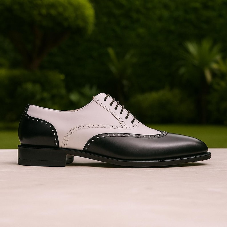 Puede incluir: Zapato Oxford de cuero de dos tonos. El zapato presenta una puntera y un tal&oacute;n negros, con una parte superior blanca. Tiene un dise&ntilde;o cl&aacute;sico con cordones y un tac&oacute;n bajo. El zapato est&aacute; sobre un fondo verde borroso.