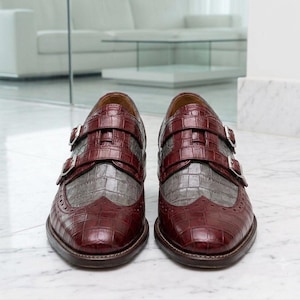 K&ouml;nnte beinhalten: Ein Paar Doppel-Monk-Schuhe in einem zweifarbigen Design aus dunkelburgunderfarbenem und grauem Leder mit Krokodilpr&auml;gung. Die Schuhe verf&uuml;gen &uuml;ber silberfarbene Metallschnallen an den Riemen und dekorative Brogue-Details an der Schuhspitze.