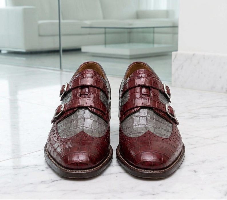K&ouml;nnte beinhalten: Ein Paar Doppel-Monk-Schuhe in einem zweifarbigen Design aus dunkelburgunderfarbenem und grauem Leder mit Krokodilpr&auml;gung. Die Schuhe verf&uuml;gen &uuml;ber silberfarbene Metallschnallen an den Riemen und dekorative Brogue-Details an der Schuhspitze.