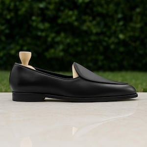 Op de afbeelding: Een zwarte leren loafer-schoen met een ronde neus en een lage hak. De schoen wordt getoond op een licht oppervlak met een groene achtergrond. Een houten schoenspanner zit in de schoen.