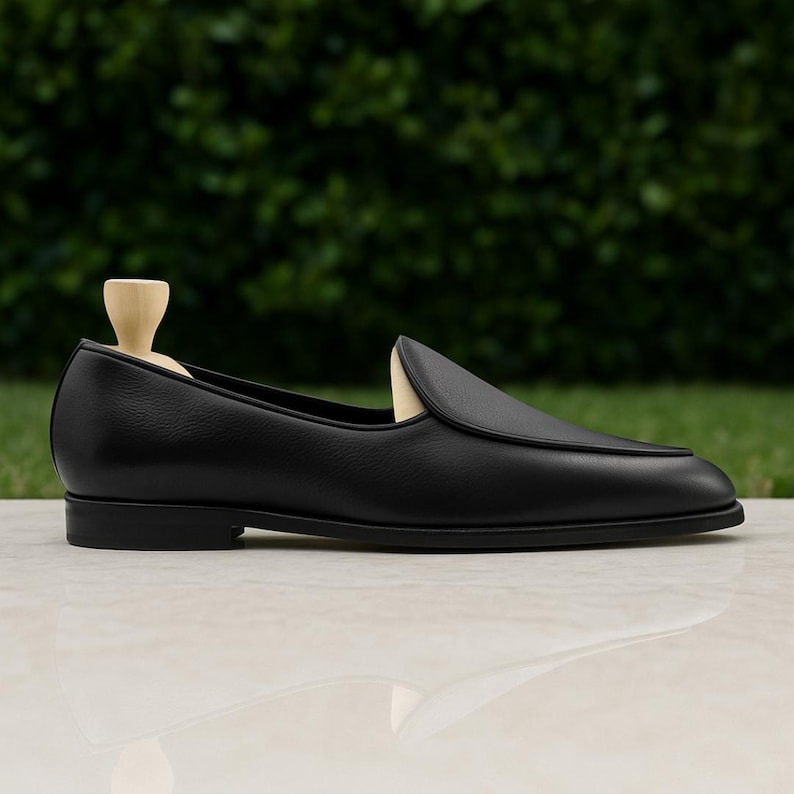 Op de afbeelding: Een zwarte leren loafer-schoen met een ronde neus en een lage hak. De schoen wordt getoond op een licht oppervlak met een groene achtergrond. Een houten schoenspanner zit in de schoen.