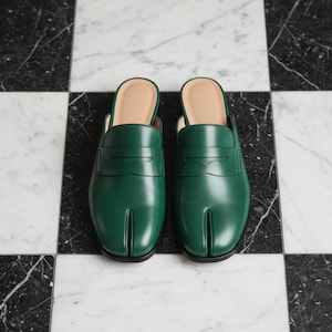 May include: A pair of green leather slip-on shoes with a split-toe design. The shoes have a light tan interior and are set against a black and white checkered marble floor. The shoes are a solid, vibrant green.