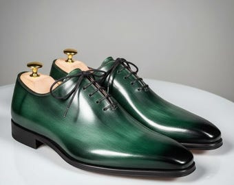 May include: A pair of green leather dress shoes with black laces. The shoes have a gradient effect, transitioning from a lighter green to a darker green at the toe. The shoes are on a white surface.