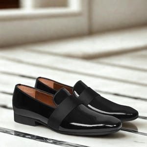 Puede incluir: Un par de mocasines negros de charol. Los zapatos tienen un acabado brillante y una correa en la parte superior. Las suelas son negras y los zapatos están sobre una superficie de mármol. Diseño clásico.