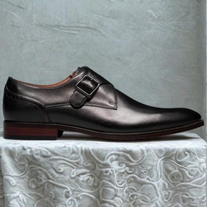 May include: A black leather dress shoe with a single buckle closure. The shoe has a dark brown sole and heel, and is displayed on a white surface.