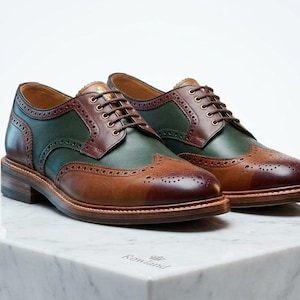 May include: A pair of leather brogue shoes featuring a multi-tone design with dark green side panels, tan toe caps, and dark brown accents. The shoes have a classic lace-up closure, decorative perforations along the seams, and a low heel.